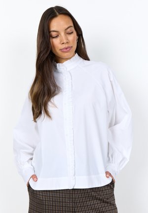 Woman with long dark hair wearing a white ruffled button-up blouse and checkered brown pants with hands in pockets.