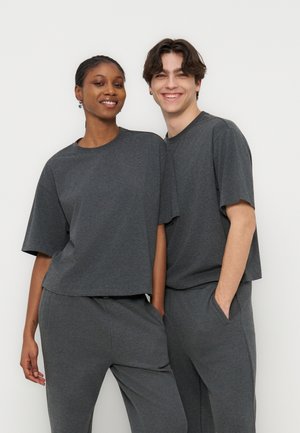 Due giovani adulti sorridenti, indossano t-shirt e pantaloni coordinati grigio antracite a vestibilità comoda, in piedi davanti a uno sfondo chiaro e uniforme.