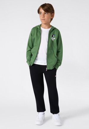Sudadera verde con cremallera, con bolsillos frontales y un diseño de caricatura en el pecho, combinada con pantalones de chándal negros y zapatillas blancas. Camiseta blanca debajo.