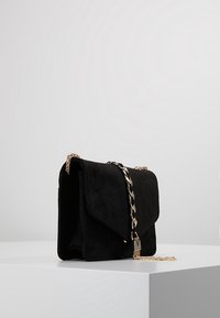Borsa a tracolla in suede nero con chiusura a pattina, tracolla in catena dorata e un accento logo metallizzato. Presenta una forma rettangolare elegante e una texture morbida.
