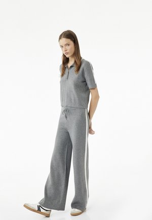 Jonge vrouw in een grijs poloshirt met korte mouwen en wijde broek met witte zijstrepen, draagt witte sneakers en staat voor een witte achtergrond.