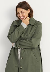 Olijfgroene trenchcoat met een klassiek ontwerp, grote knopen, zijzakken en een brede kraag, gedragen over een lichtgrijze trui.