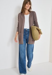 Cárdigan marrón de punto con frente abierto y bolsillos, combinado con jeans de pierna ancha azul claros y zapatillas blancas, sosteniendo un bolso acolchado verde oliva.