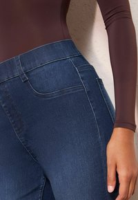 Jeans de mezclilla azul oscuro con una textura suave, cintura alta y un sutil detalle de costura, combinados con una blusa ceñida de manga larga marrón.