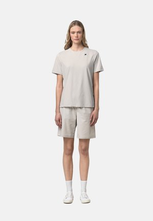 Donna in piedi, rivolta in avanti, che indossa una maglietta oversize beige chiaro e pantaloncini coordinati con calzini bianchi e scarpe da ginnastica su uno sfondo semplice.