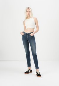 Vysoké skinny džínsy v tmavomodrom denime s hladkou textúrou, skombinované s priliehavým bielym crop topom a čiernymi teniskami s béžovými akcentmi.