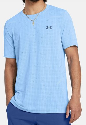 Mężczyzna ubrany w jasnoniebieską koszulkę Under Armour z krótkim rękawem z subtelnym wzorem w kratkę, niebieskie spodnie, złoty naszyjnik z krzyżem i widoczny tatuaż na lewym przedramieniu.