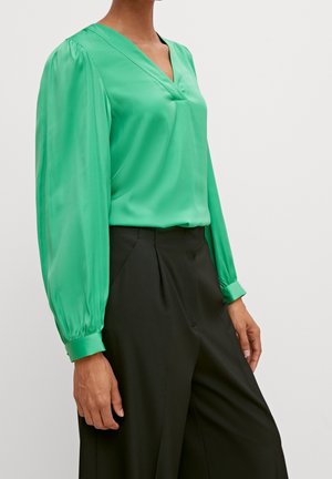 Kvinde iført en lysegrøn bluse med lange ærmer og puffede ærmer samt sorte højttaljede bukser mod en ensfarvet baggrund.