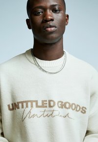 Jonge zwarte man in een witte trui met de tekst "UNTITLED GOODS" en twee zilveren kettingen tegen een effen lichte achtergrond.