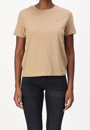 Osoba ubrana w beżowy t-shirt z krótkim rękawem i czarne jeansy, pokazana od ramion do bioder na jednolitym tle.