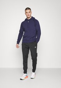 Marineblauwe hoodie met een voorzak, gedragen met zwarte tapers joggingsbroeken met zijaccenten, en witte sneakers met kleurrijke details.