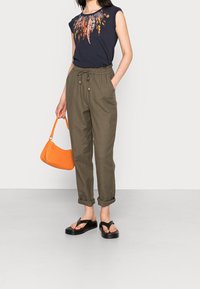 Vrouw in een zwart top met verenpatroon, olijfgroene broek met trekkoord, opgerold aan de mouwen, zwarte platform sandalen, met een oranje handtas.