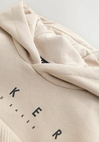 Béžová mikina s textúrovanou tkaninou, s veľkým čiernym potlačeným logom a kapucňou so šnúrkou. Detailný záber ukazuje švy.