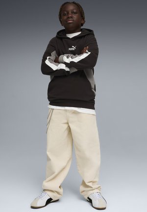 Junge Person steht selbstbewusst mit verschränkten Armen, trägt schwarzen und grauen Puma-Hoodie, beige weit geschnittene Hose und weiße Sneakers.