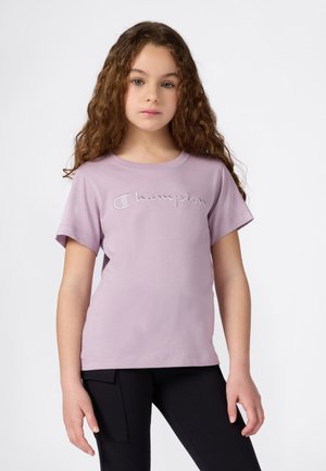 Fille aux cheveux bouclés portant un t-shirt Champion violet clair et un pantalon noir, se tenant devant un fond blanc uni.