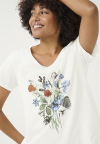 T-shirt en coton blanc avec un col en V, ornée d'un imprimé floral coloré en rouge, bleu et vert, représentant diverses fleurs et feuilles.
