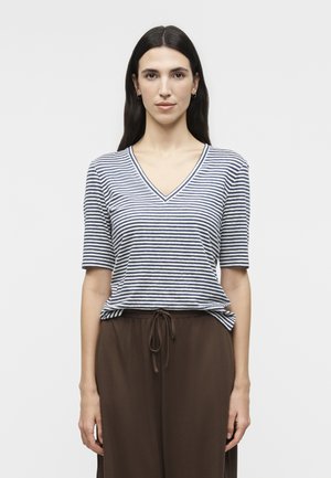 Γυναίκα με μακριά σκούρα μαλλιά, που φοράει κρίκους στα αυτιά, μπλε και λευκή ριγέ μπλούζα με V λαιμόκοψη και καφέ παντελόνι με κορδόνι, που στέκεται μπροστά σε λευκό φόντο.