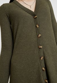 Cardigan côtelé vert olive avec un col en V, cinq boutons marron sur le devant, et un tissu en tricot texturé. Manches longues.