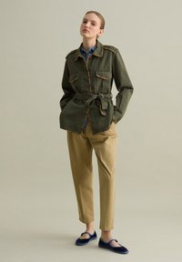 Giacca verde oliva con ricami colorati lungo il colletto e le tasche, vita con cintura, abbinata a pantaloni beige e ballerine blu navy.