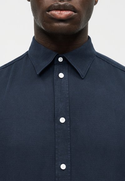 Mężczyzna o ciemnej karnacji ubrany w granatową koszulę zapinaną na guziki z kołnierzykiem na spinki na jednolitym jasnym tle.