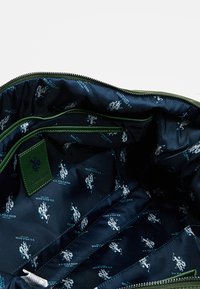 Zielona torba na zamek błyskawiczny z ciemnoniebieskim wnętrzem, ozdobiona powtarzającym się wzorem logo U.S. Polo Assn. Gładka tkanina z kontrastowymi akcentami i kieszeniami.