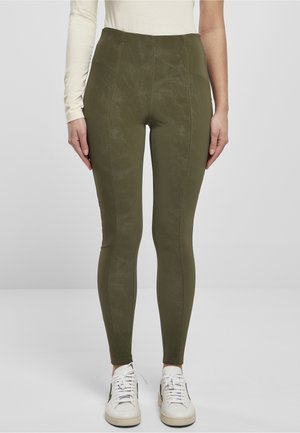 Kvinde iført olivengrønne leggings med højt liv og synlige sømme, kombineret med hvide sneakers og en beige langærmet top på en ensfarvet baggrund.