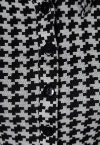 Svart-vit houndstooth-mönstrad stickad tyg med sex svarta knappar framtill. Slät textur och medelhög tjocklek.