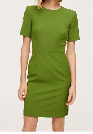 Kobieta ubrana w dopasowaną zieloną sukienkę do kolan z krótkimi rękawami i okrągłym dekoltem, stojąca na tle jednolitego jasnego tła.