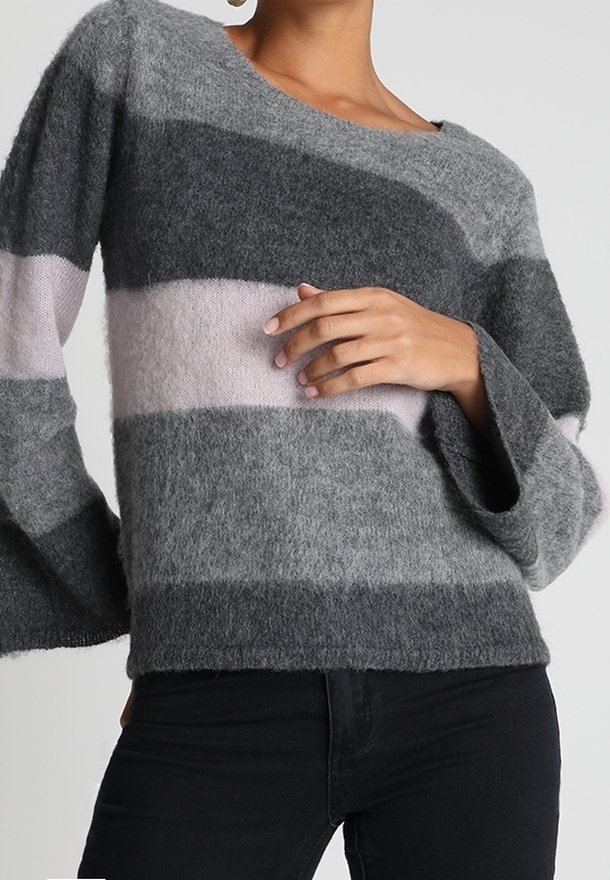 Femme portant un pull gris duveteux avec de larges rayures horizontales en gris foncé, rose clair et gris moyen, associé à un pantalon noir.
