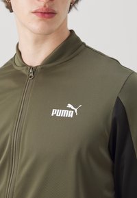 Maslinasto zelena zip-up jakna s crnim detaljima; karakterizira je glatka tekstura i bijeli Puma logo na prsima. Visoki ovratnik.