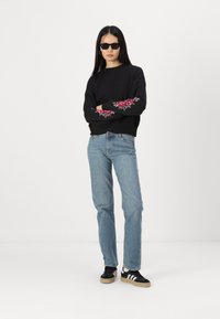 Μαύρη μπλούζα με ροζ λουλουδάτη κεντητική στα μανίκια, σε συνδυασμό με ανοιχτό γαλάζιο τζιν και μαύρα αθλητικά παπούτσια με άσπρες ρίγες.