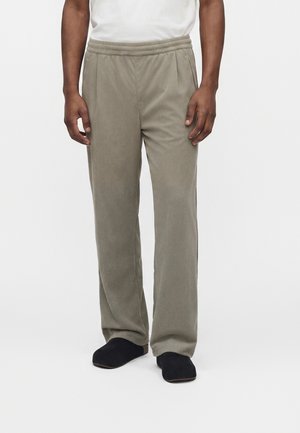 Persona che indossa pantaloni di velluto beige con vita elastica e scarpe slip-on nere, in piedi su uno sfondo bianco semplice.