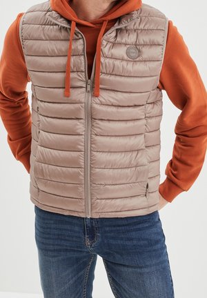 Homme portant une veste matelassée marron clair par-dessus un sweat à capuche orange et un jean bleu, les mains dans les poches, sur fond blanc.