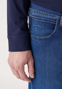 Camisa de manga larga azul marino con puños acanalados, combinada con unos vaqueros de mezclilla azul que presentan costuras naranjas contrastantes y un bolsillo frontal.