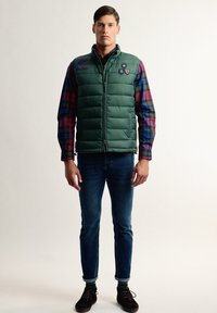 Chaleco puffy verde con cuello alto, cierre de cremallera y parches; superpuesto a una camisa de cuadros multicolor; combinado con jeans azules y zapatos marrones.