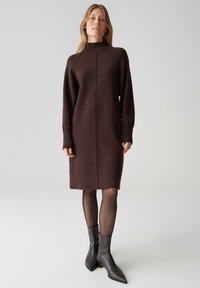 Bruine gebreide jurk met een hoge hals, lange mouwen en een rechte snit. Voorzien van geribde manchetten en een centrale naaddetail.