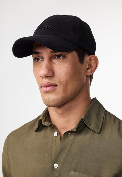 Black cotton cap with curved brim and stitched logo "NN. 07." Model wears a green shirt with buttoned collar and textured fabric.