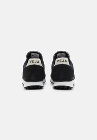 Black suede sneakers with a white rubber sole, featuring a gray heel tab and “VEJA” branding on the heel in cream-colored text.