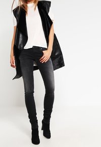 Black sleeveless vest with glossy and textured fabric, worn over a white T-shirt, paired with black skinny jeans and ankle boots.