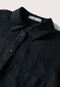 Camicia in lino nera con colletto, taschino frontale e bottoni neri. Il tessuto presenta una finitura testurizzata e una caduta morbida.
