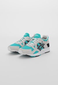 Ett par teal-, grå- och svarta Asics Gel-Lyte V-sneakers med vita snörningar och sulor, vinklade lite åt vänster mot en vit bakgrund.