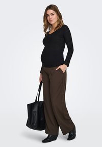 Schwarzes, geripptes Langarmoberteil, braune Hosen mit weitem Bein und schwarze Keilstiefel. In einer Hand hält das Model eine schwarze Tragetasche. Das Model hat mittellanges Haar.