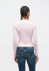 Persoon met gevlochten haar draagt een lichtroze top met lange mouwen en blauwe spijkerbroek, staand met het gezicht naar een effen witte achtergrond.