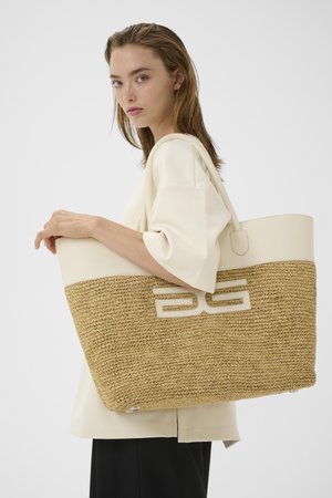 Kvinde med lyst hår iført en cremefarvet bluse holder en stor flettet stråtaske med cremefarvet lædertop og håndtag.