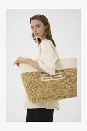 Kvinde med lyst hår iført en cremefarvet bluse holder en stor flettet stråtaske med cremefarvet lædertop og håndtag.