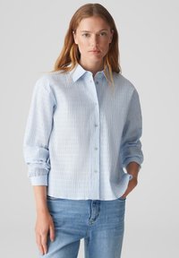 Camicia button-up azzurro chiaro, stropicciata, con colletto, maniche lunghe e vestibilità rilassata. Presenta sottili righe verticali e una texture liscia.