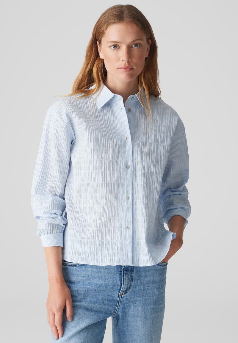 Camicia button-up azzurro chiaro, stropicciata, con colletto, maniche lunghe e vestibilità rilassata. Presenta sottili righe verticali e una texture liscia.