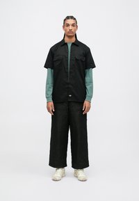 Černá košile s krátkým rukávem a knoflíky s dvěma kapsami, přes ní je navlečená dlouhá tyrkysová košile. Volné černé prošívané kalhoty a béžové tenisky.