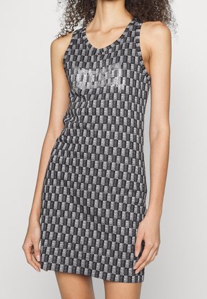 Robe sans manches à carreaux noirs et gris, silhouette ajustée avec un logo métallique « Guess » sur la poitrine, portée par une personne debout.