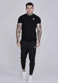 Čierne priliehavé tričko s krátkym rukávom a malým logom, skombinované s čiernymi cargo teplákmi s bočnými vreckami, nosené s bielymi teniskami.